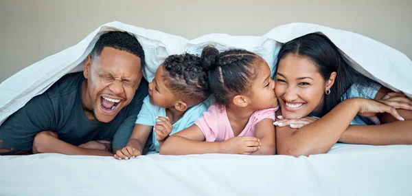 Black family, bed together and happy under blanket in home with love, care and bonding with kiss on cheek. Black woman, man and kids in bedroom in house for happy family moment, time or relax in home.