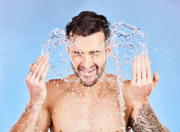 Face, water and cleaning with a man model splash in studio on a blue background for hygiene. Skincare, water splash and hydrate with a handsome young male washing his skin in the bathroom for care.