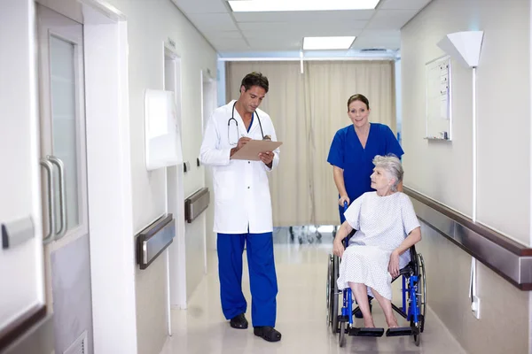 あなたは本当によくやっています 医者と話をしながら廊下を車椅子で老人を押している女性看護師 — ストック写真
