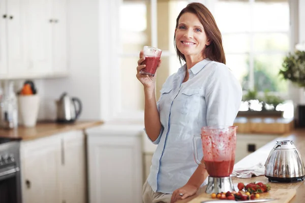 This beats coffee any day. An attractive brunette enjoying a fruit smoothie while leaning against a kitchen counter