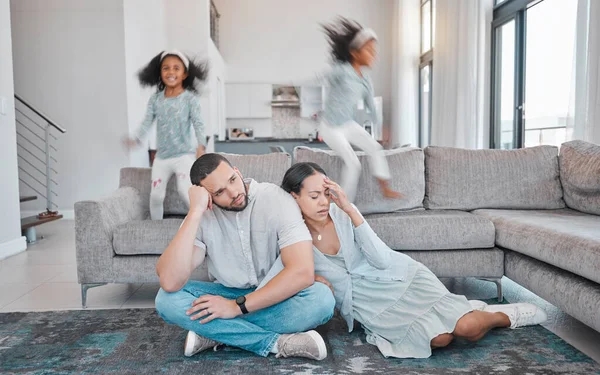 Tired, parents and children jump on sofa with frustrated mom and dad sitting on floor in living room. Family, exhausted couple and playful kids with energy jumping on couch at home during quarantine.