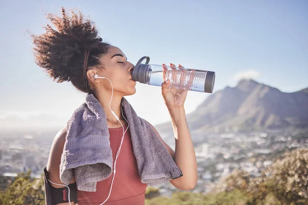Music, workout and woman drinking water after exercise at sunrise along mountain, wellness and training on blue sky. Radio, water and black woman stop for break from fitness, running and cardio run.