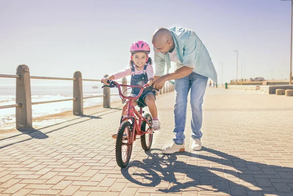 Father, child and bicycle with a girl learning to ride a bike on promenade by sea for fun, bonding and quality time on summer vacation. Man teaching his daughter or girl safety while cycling outdoor.
