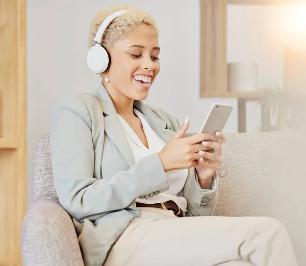 Business woman, headphones and phone streaming of music or video in a office with technology. Black woman smile on social network, music or web app on a sofa listening to funny podcast.