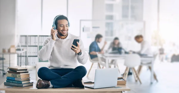 Man, music headphones and relax at startup, office or workplace for digital marketing with smile. Happy worker, video and sitting on table with smartphone, laptop and reading social media at company.