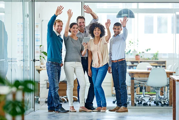 This team goes hand in hand with success. Portrait of a diverse group of coworkers waving at the camera while standing in an office