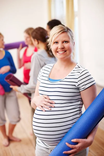 Keeping in shape during pregnancy. A young blonde pregnant woman smiling while holding her exercise mat while her friends stand in the background