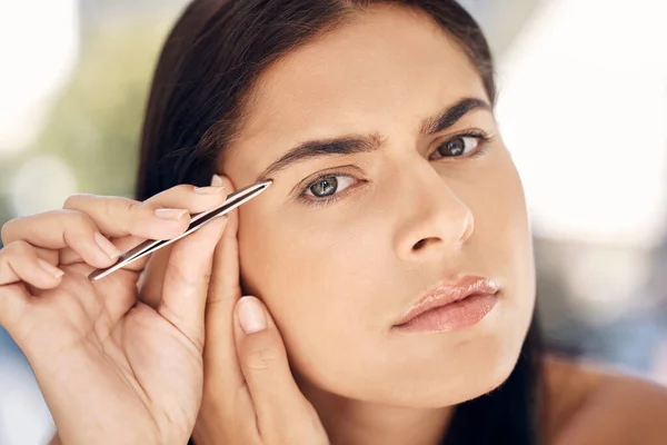 Woman in mirror plucking eyebrows with tweezers and grooming facial routine in morning. Beauty, skincare and tweezing eyebrow hair, portrait of Indian woman face in reflection in bathroom treatment