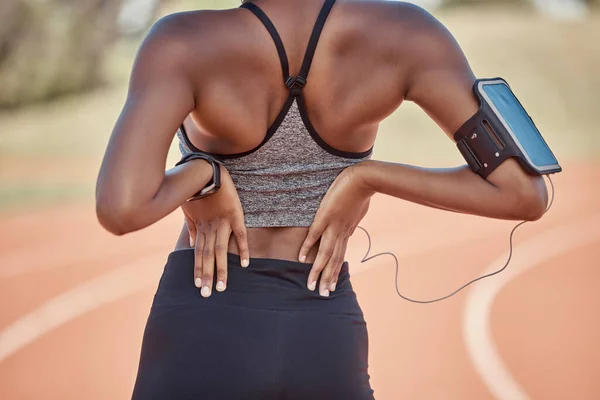 Running, woman on track with back pain and phone for fitness app on outdoor workout. Technology, sports injury and black woman with smartphone on digital app streaming music for health and wellness