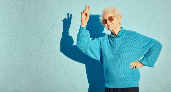 青い背景の壁に平和のサインを持つ都市 ファッション シニア女性は 週末と退職をお楽しみください エネルギー 手のサインと町のクールでモダンな流行の服を持つ幸せな高齢者の女性 — ストック写真
