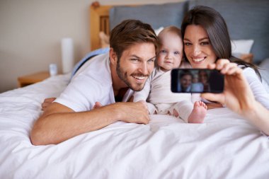 Aile özçekimi. Küçük kızlarıyla yatakta uzanan ve cep telefonlarıyla selfie çeken genç bir çift.