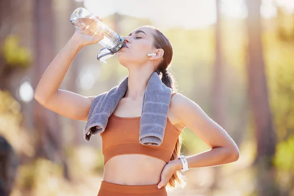 Fitness, woman and drinking water after running, training and cardio exercise in a forest. Sports, water and girl drinking after run in nature with music, podcast or radio for audio track by runner.