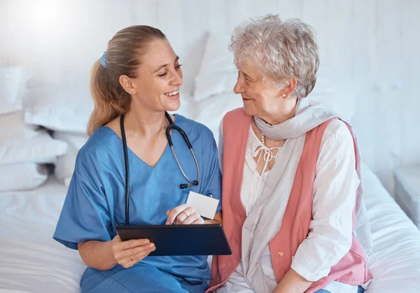 Happy, tablet and nurse consulting an old woman with medical report results or doctor healthcare diagnosis. Smile, bed and social worker helping, talking or speaking to a healthy elderly person.