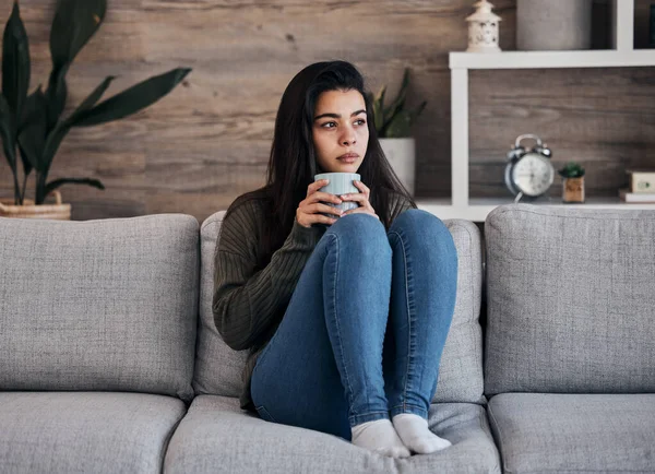 Depression, sad and woman with coffee on a sofa for thinking, lonely and isolated in a living room. Stress, anxiety and girl drinking tea on couch, looking depressed about debt or mental health.