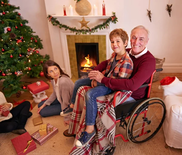 Family, christmas and love of child, grandpa and mother together to open gift or present together at fireplace in home living room. Senior man in wheelchair with boy and woman to celebrate holiday.