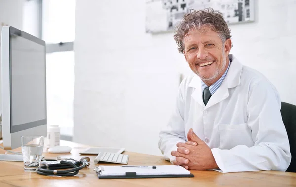 总是乐于帮忙的一个面带微笑的成熟医生坐在办公室桌子旁的画像 — 图库照片