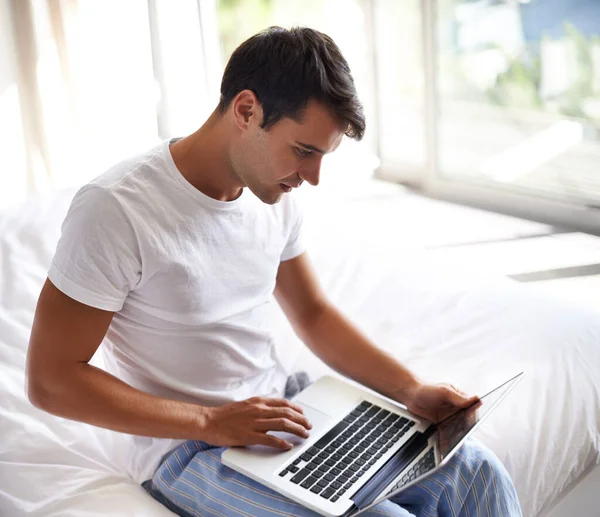 在早上查他的电子邮件一个英俊的年轻人在床边用笔记本电脑 — 图库照片