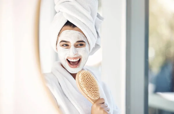 Woman, face mask and mirror with a brush for singing, beauty and home spa treatment in bathroom with a towel and bathrobe. Face of happy female doing facial for self care and detox with cosmetics.