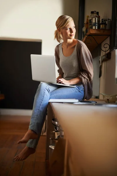 从她厨房的舒适感开始写博客 一位年轻貌美的女士在厨房里轻松地使用笔记本电脑 — 图库照片