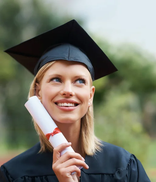 成功は彼女の顔に笑顔を置きます 遠くを見ながら帽子とガウンを身に着けている若い大学の卒業生と笑顔 — ストック写真