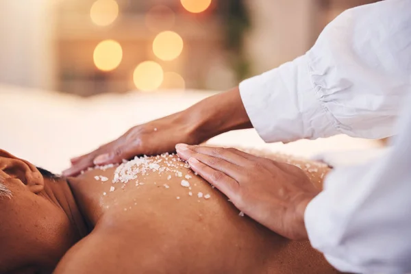Spa, massage and salt on back for detox, exfoliate and skin care with hand of therapist for luxury treatment for health and wellness. Client on table to relax with body scrub cosmetic at beauty salon.