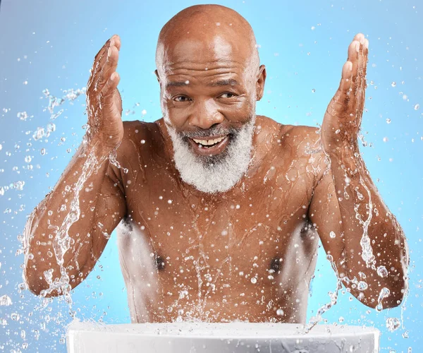 Water splash, senior and black man cleaning face in studio isolated on a blue background. Skincare, hygiene and retired elderly male from Nigeria bathing or washing for wellness and healthy skin
