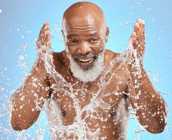 Black man cleaning face, water splash and skincare with beauty, hygiene and facial care in portrait against blue background. Mature model with water, splash and skin hydration, wellness and grooming
