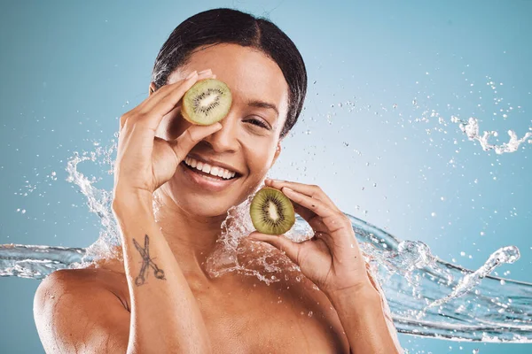 Skincare, water and portrait of woman with kiwi for natural, organic and healthy beauty products in studio. Dermatology, wellness and girl with fruit, water splash and cosmetics on blue background.