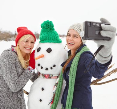 Selfie, kardan adam ve kadın arkadaşlarla dışarıda karda fotoğraf çektirmek. Mutlu, gülümseyen ve fotoğraf çeken genç bir bayan ve arkadaşı soğuk havada fotoğraf için poz veriyor..