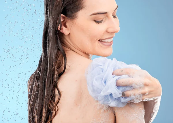 Skincare, back cleaning and woman in shower in studio isolated on a blue background. Face, water splash and female model from Canada bathing or washing with sponge for beauty, body care or wellness