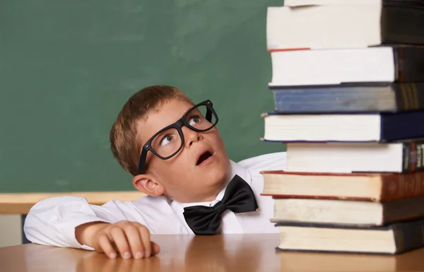 Who said school is easy. A young boy wearing glasses and a bow-tie looking wearily at a large pile of books