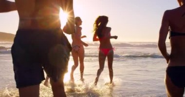 Arkadaşlar, plaj ve insanlar eğlence olsun diye suda koşarken gün batımında bikinili ve açık havada mayo giyiyorlar. Erkekler ve kadınlar yaz tatilinde okyanus suyunda bir araya gelirler..