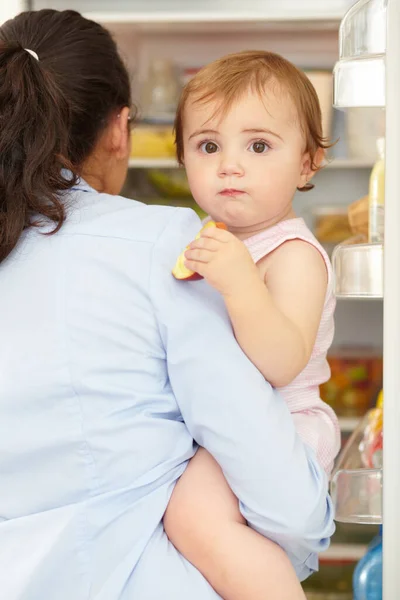 Im one hungry baby. a cute baby being held by its caring mom in front of an open fridge