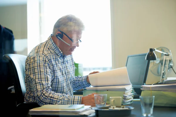 经验丰富 效率高 一个高级商人戴着耳机在办公桌前办理文书工作 — 图库照片