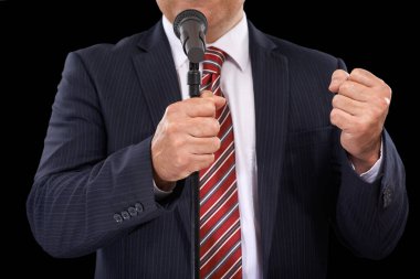 O, söylediğine inanmaktadır. Takım elbiseli bir adamın mikrofona yumruklarıyla konuşmasının görüntüsü.
