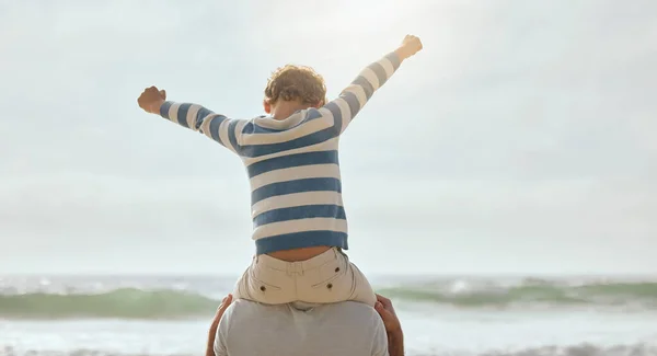 Cute little caucasian boy from behind cheering with arms outstretched while sitting on his fathers shoulders at the beach. Playful young child from the back having fun and bonding with dad outdoors. .