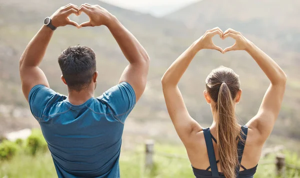 Fit young man and woman from the back gesturing heart shapes with hands while exercising together outdoors. Two athletes caring for body with regular training workout or run. Endorsing and loving a h.