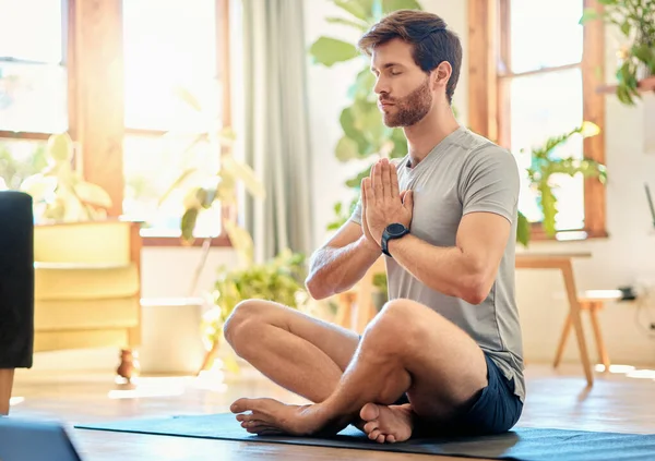 One young caucasian man sitting with legs crossed and eyes closed meditating in harmony with hands together in namaste gesture while practising yoga at home. Calm, relaxed and focused guy feeling zen.