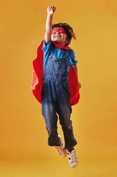 Adorable little asian boy wearing a mask and a cape while pretending to be a superhero against an orange studio background. Cute happy boy pretending to be a character for halloween.