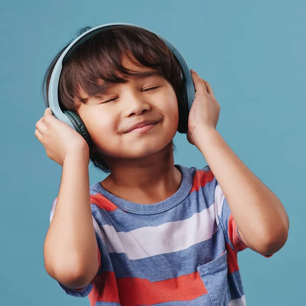 A cute young asian boy enjoying listening to music from his wireless headphones. Adorable Chinese kid smiling and feeling the magic of music while posing against an blue studio background.