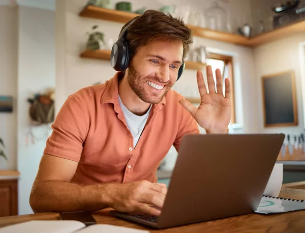 Young happy caucasian businessman wearing headphones and waving his hand on a virtual meeting on a laptop at home alone. One joyful male businessperson smiling while working in the kitchen at home.