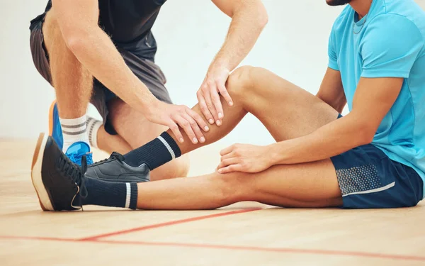 Unknown athletic squash player helping friend with injury after playing game on court. Mixed race athlete suffering from shin splints during training practice at sports centre. Physio massage for pai.