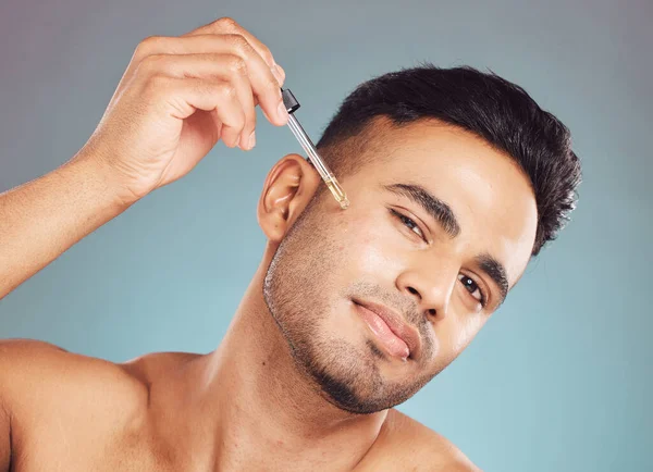 Portrait of one handsome young indian man using a dropper to apply serum oil to his skin and face against a blue studio background. Mixed race guy using a moisturising aftershave product for healthy,.