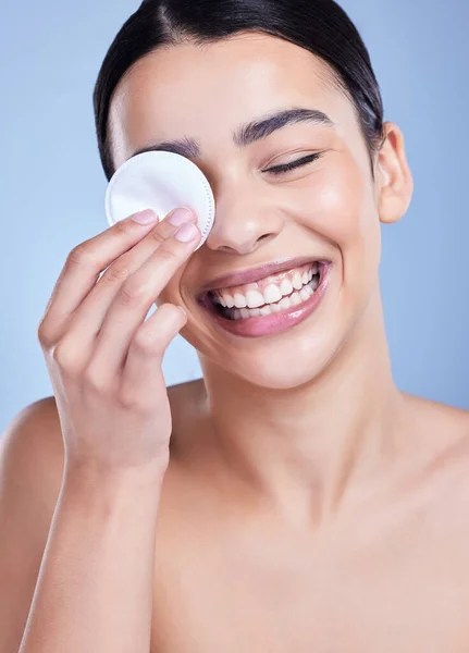 A beautiful smiling mixed race woman using a cotton pad to remove makeup during a selfcare grooming routine. Hispanic woman applying cleanser to her face against blue copyspace background.
