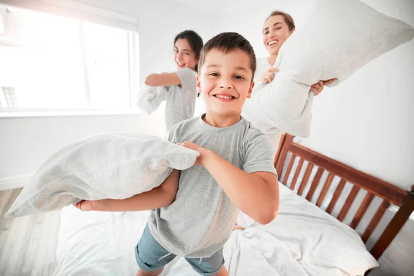 Portrait of adorable little boy, sister and mother having a fun pillow fight at home. Happy young family with mother and two kids holding pillows and standing on bed playing and enjoying free time to.