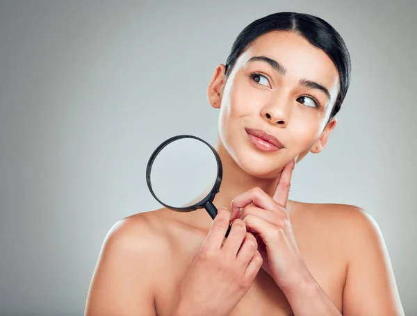 A beautiful mixed race woman posing with a magnifying glass. Young hispanic obsessed with targeting acne and wrinkles against a grey copyspace background.