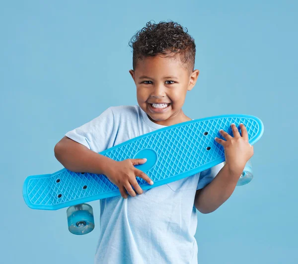 Excited little hispanic boy holding his skateboard. Happy smiling kid hugging skateboard after getting it as a gift against blue studio background.