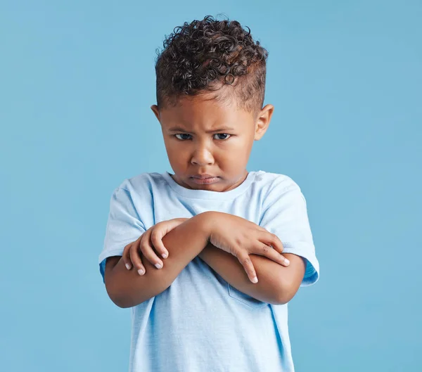 Offended little hispanic boy looking sad and upset while standing against a blue studio background. Unhappy preschooler standing with his arms crossed and looking down.