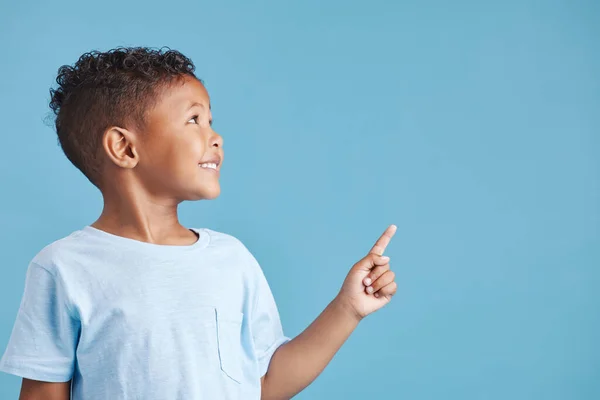 Close up of a happy smiling little boy looking up and pointing his finger at copy space to the right against blue studio background. Cheerful mixed race kid in casual clothes.