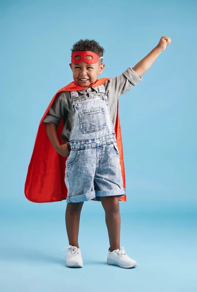 Portrait of cheerful little boy in superhero cape and mask stretching out his hand on blue background. Strong brave kid ready to save the world with his superpowers.